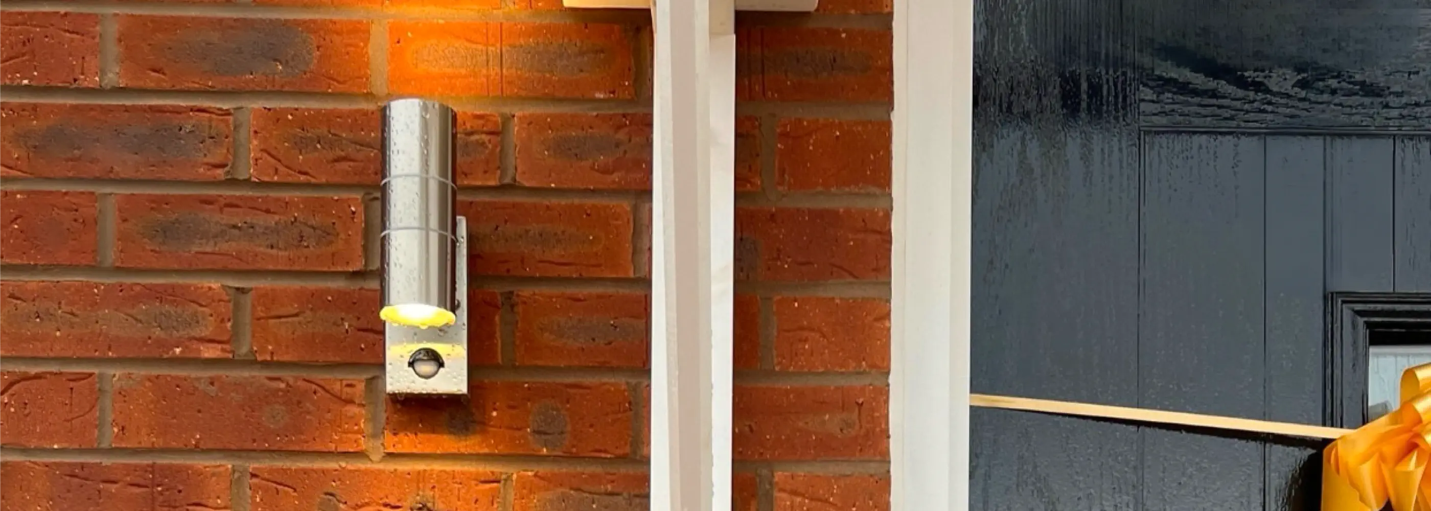 A modern house number sign displaying “No 8” in white text on a dark rectangular plaque is mounted on a red brick wall next to a sleek black door and a white vertical light fixture.