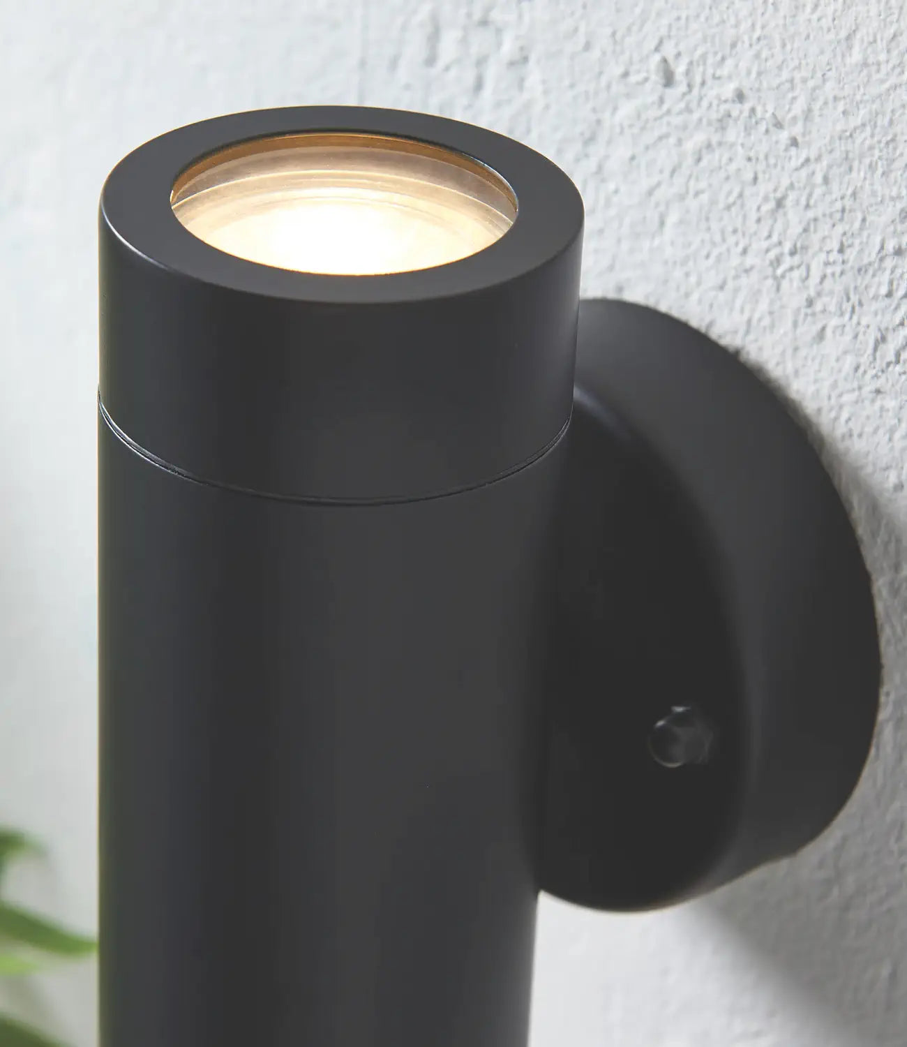 A close-up of a modern black cylindrical wall light mounted on a textured white wall, highlighting its sleek finish and warm upward illumination.