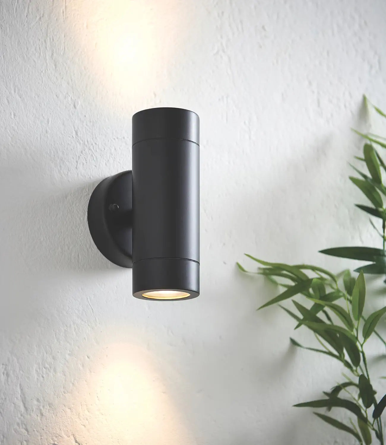 A sleek black cylindrical up-and-down outdoor wall light mounted on a white textured wall, casting warm light both upward and downward, with greenery adding a natural touch to the scene.