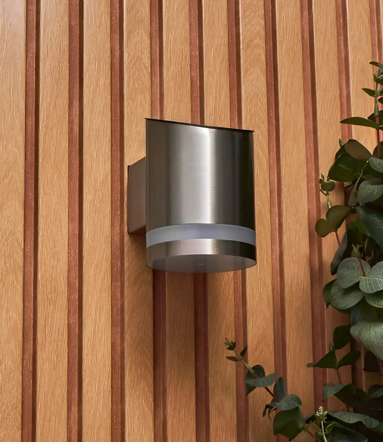 A contemporary brushed stainless steel outdoor wall light with a rounded design, mounted on a vertically slatted wooden wall, complemented by lush green foliage on the side for a natural aesthetic.