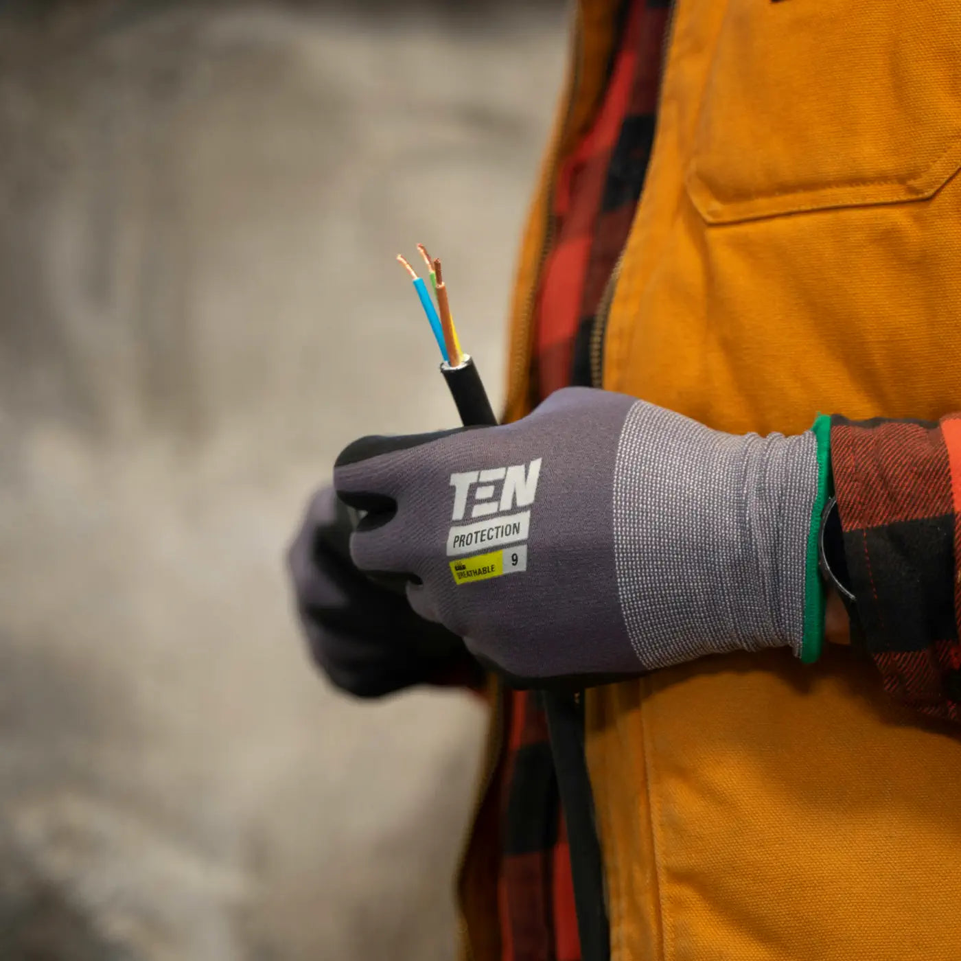 A person wearing protective gloves holds a section of electrical wiring with exposed copper ends, ready for connection. They are dressed in a yellow jacket and flannel shirt.