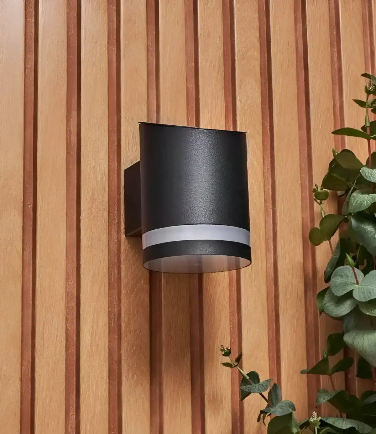 A modern black outdoor wall light with a cylindrical design, mounted on a vertically slatted wooden wall, accented by greenery on the side for a sleek and natural look.