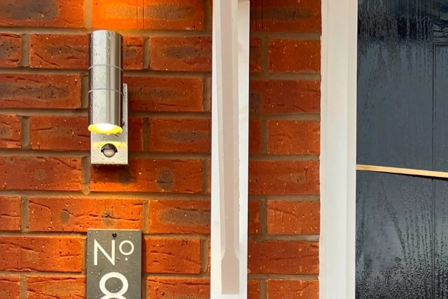 A modern house number sign displaying “No 8” in white text on a dark rectangular plaque is mounted on a red brick wall next to a sleek black door and a white vertical light fixture.
