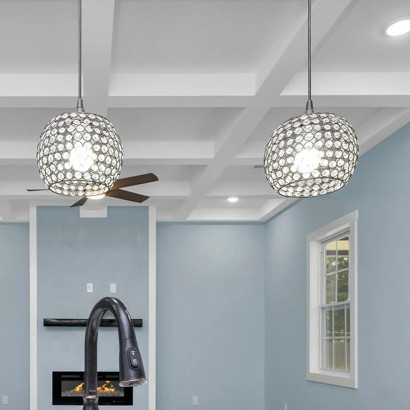 A modern kitchen with two pendant lights featuring decorative crystal-like shades, hanging from a coffered ceiling. The space includes blue walls, a window, and a sleek black faucet in the foreground.