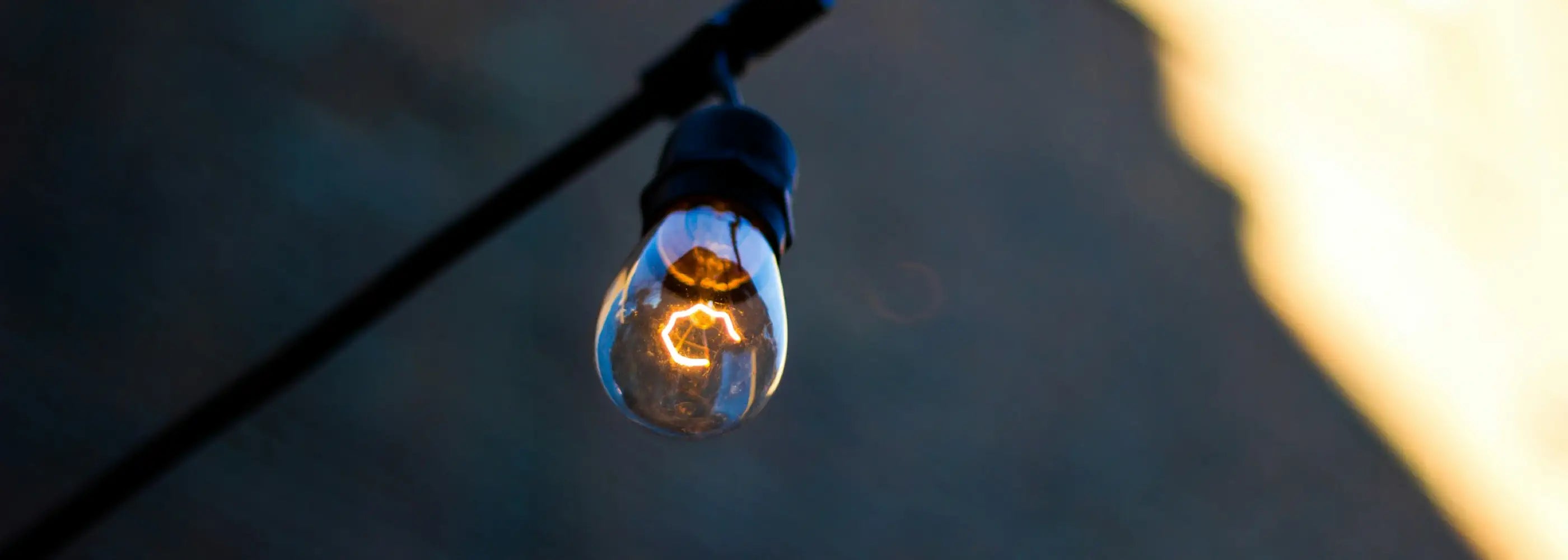 A close-up of a glowing filament bulb hanging from a black cable, set against a blurred background with soft blue and golden tones, creating a warm and moody atmosphere.