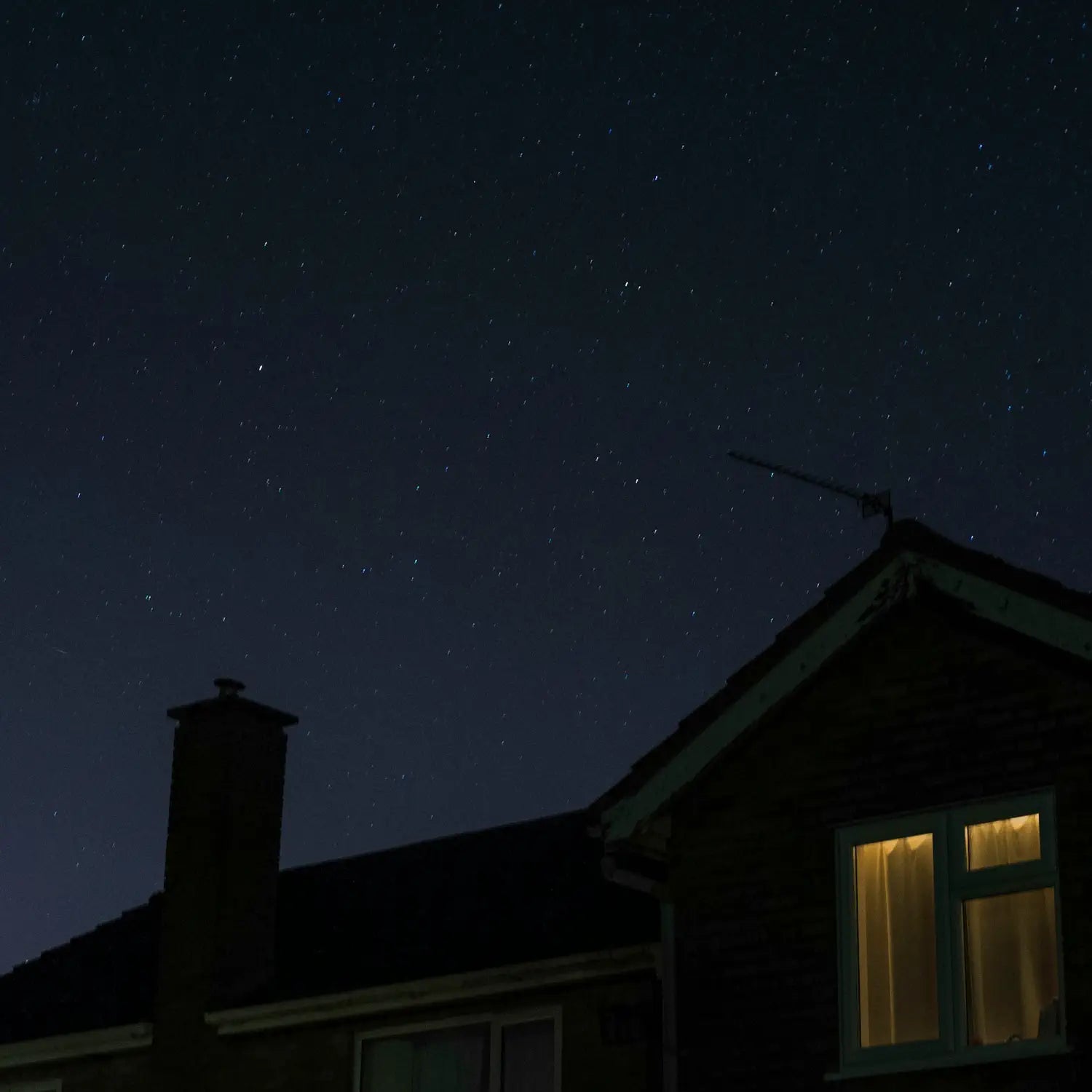 A house silhouette at night under a clear, star-filled sky, with a warm glow coming from a lit window.