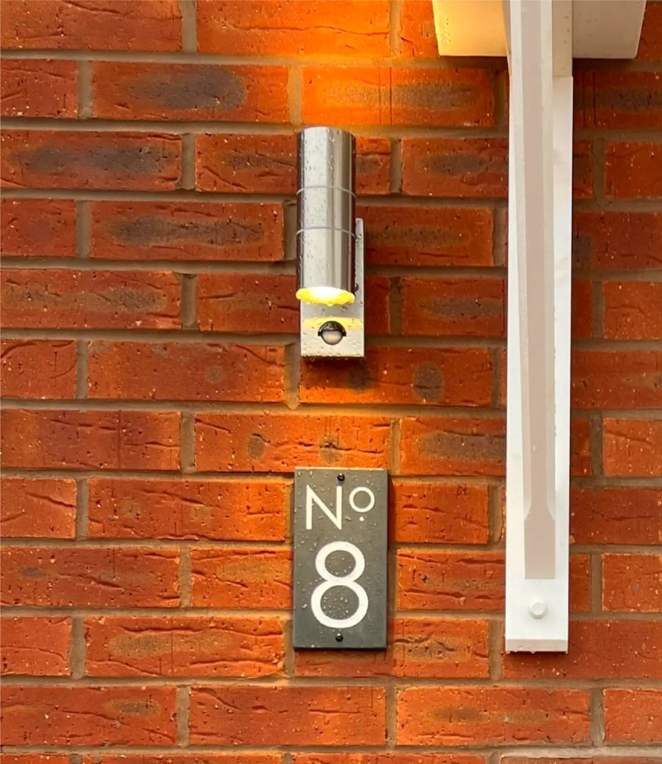 A modern house number sign displaying “No 8” in white text on a dark rectangular plaque is mounted on a red brick wall next to a sleek black door and a white vertical light fixture.