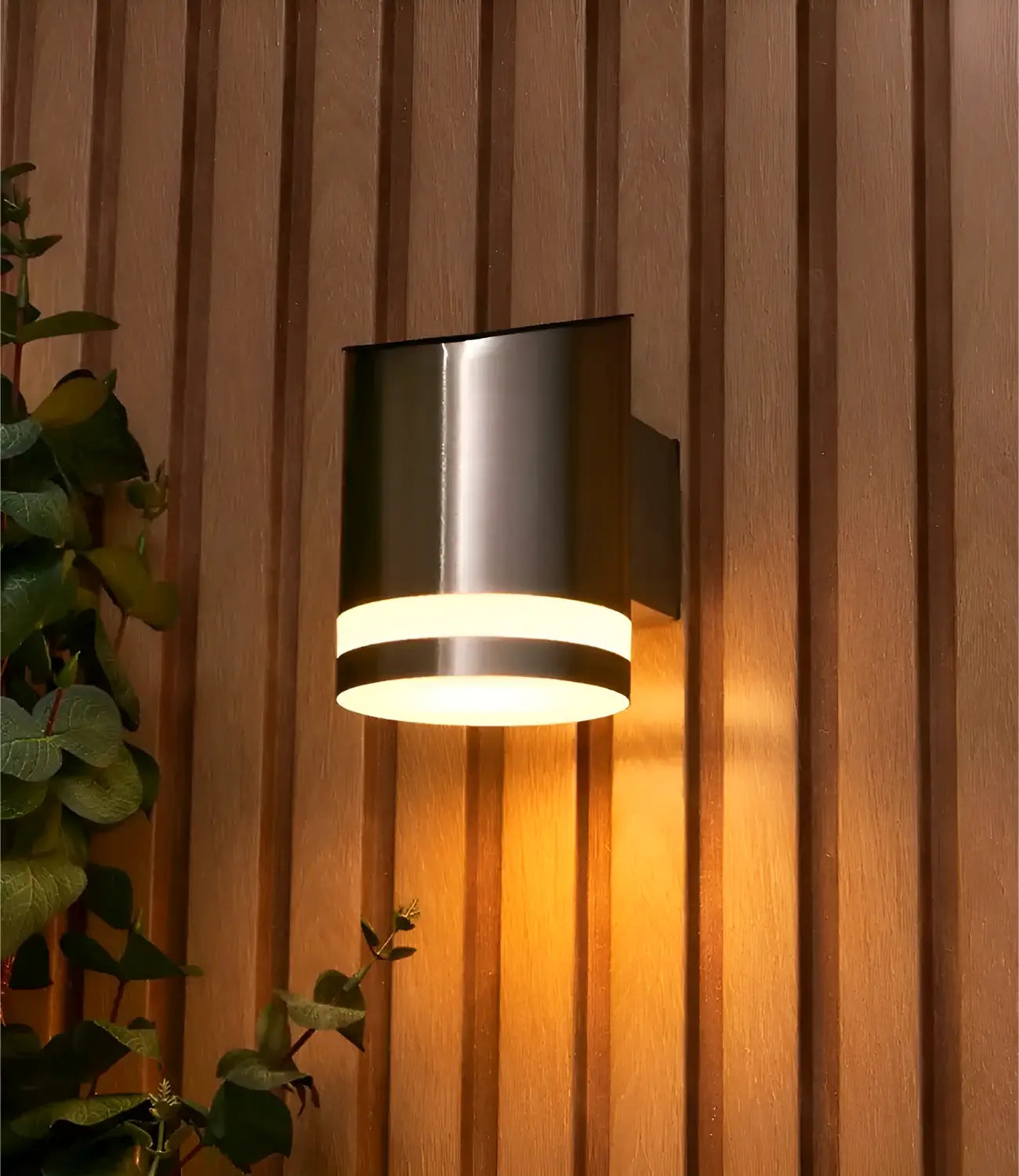 A stylish brushed stainless steel outdoor wall light illuminated with a warm glow, mounted on a vertically slatted wooden wall, accompanied by greenery on the side for a modern and inviting appearance.
