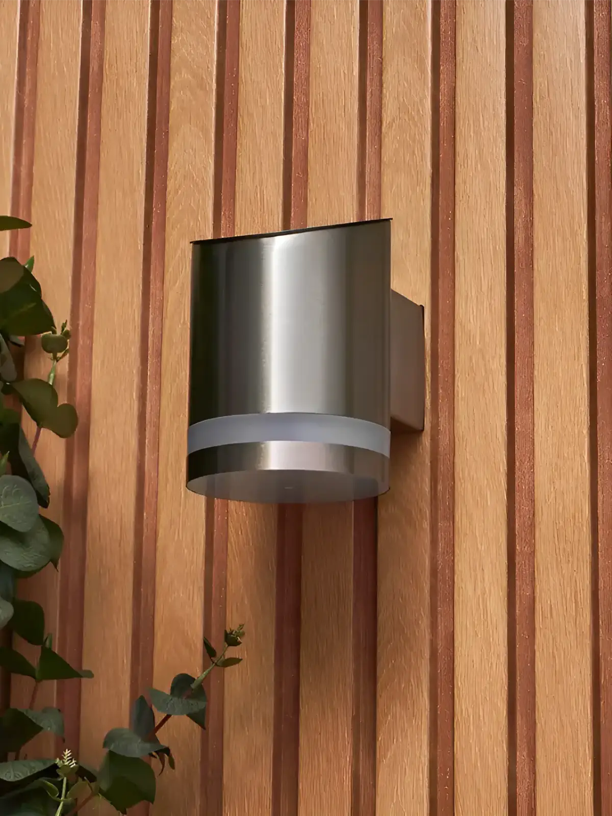 A modern brushed stainless steel outdoor wall light with a curved design, mounted on a vertical wooden panel, complemented by greenery on the side for a natural aesthetic.