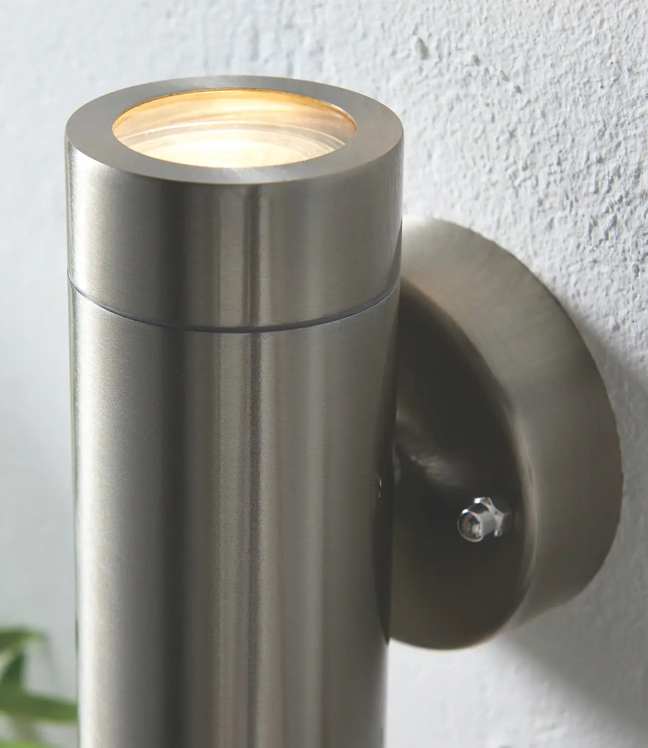 A close-up of a brushed stainless steel cylindrical wall light mounted on a textured white wall, showcasing its sleek design and warm upward illumination.