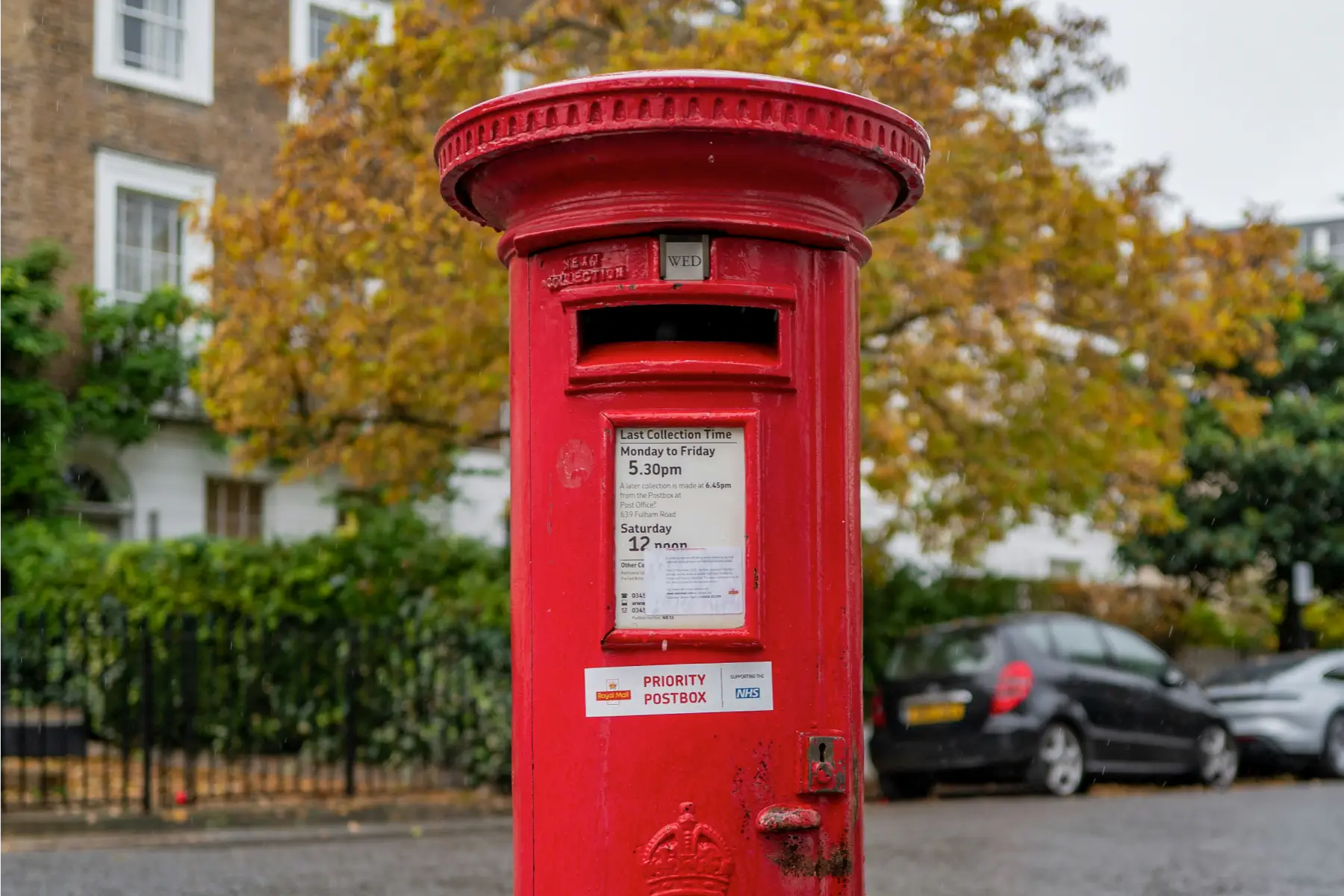Traditional red British postbox on a street with trees and parked cars in the background.