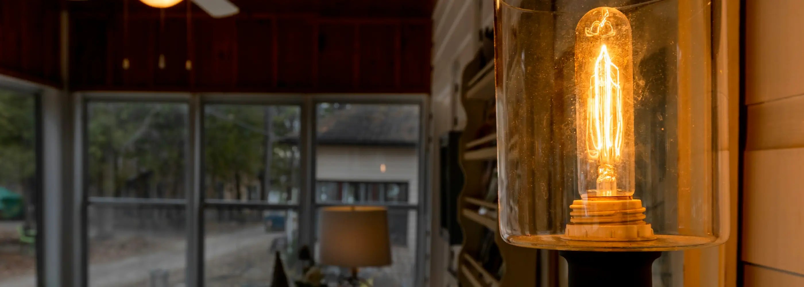 A warm filament bulb glows inside a glass sconce, illuminating a cozy interior space with wooden ceilings and large windows overlooking an outdoor area.