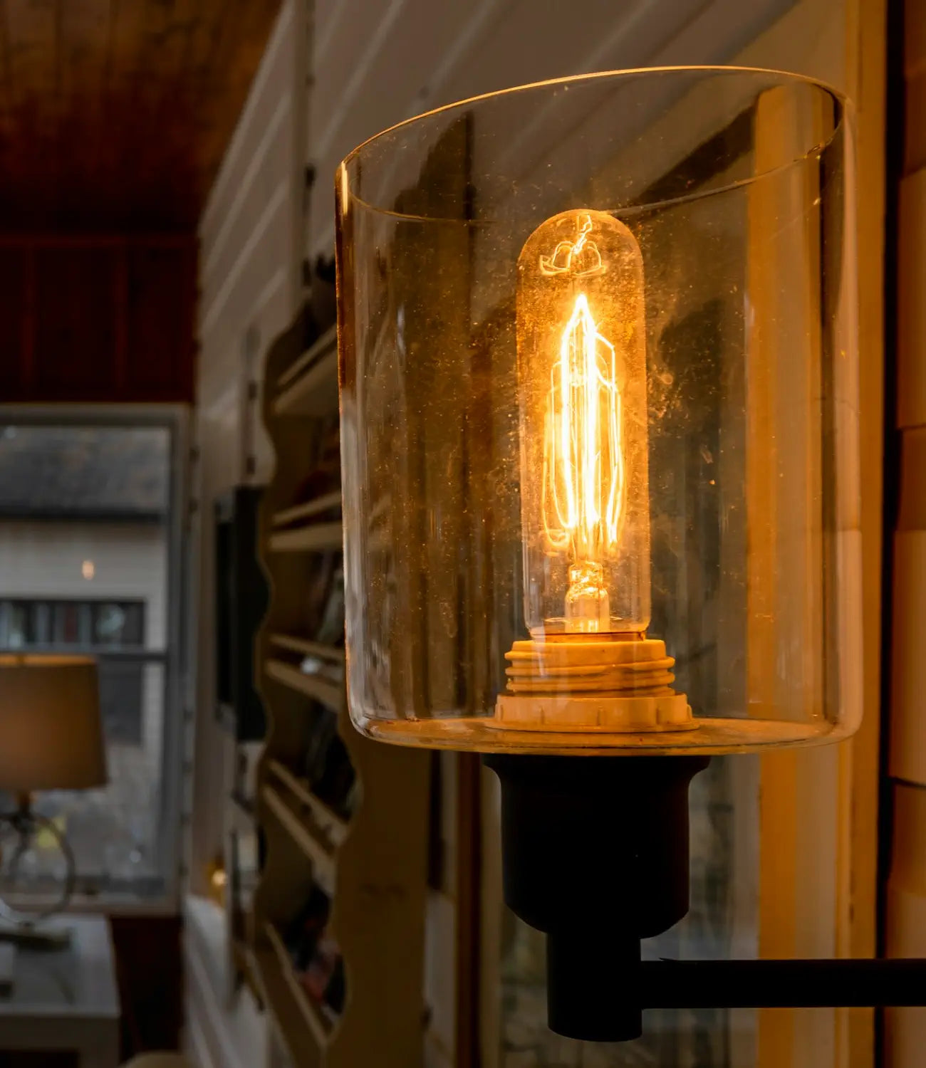 A warm filament bulb glows inside a glass sconce, illuminating a cozy interior space with wooden ceilings and large windows overlooking an outdoor area.