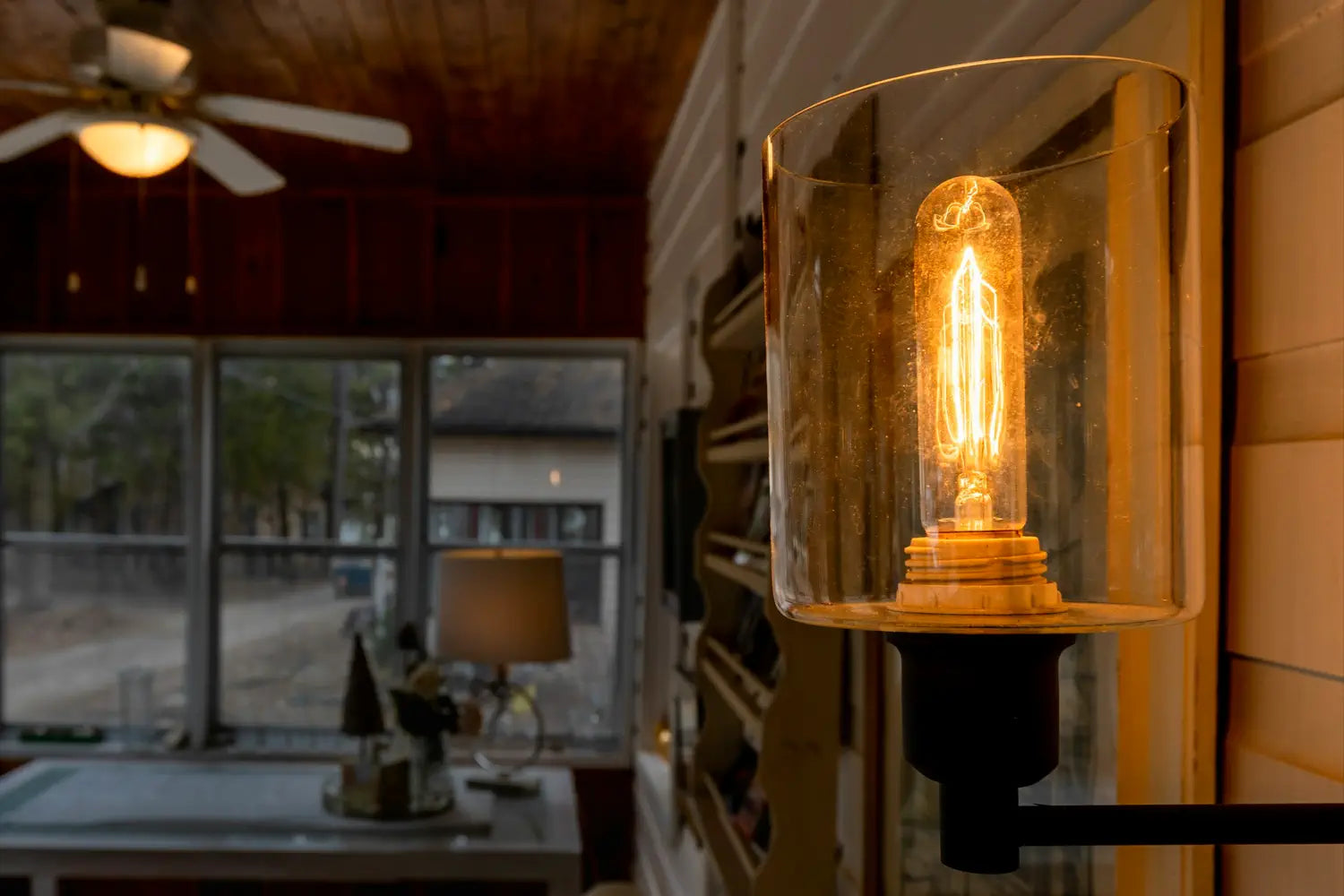 A warm filament bulb glows inside a glass sconce, illuminating a cozy interior space with wooden ceilings and large windows overlooking an outdoor area.