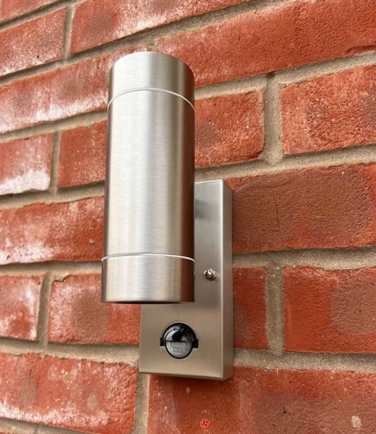 A brushed stainless steel outdoor wall light with a cylindrical design and an integrated motion sensor at the base. It is mounted on a red brick wall, blending modern functionality with a classic exterior backdrop.