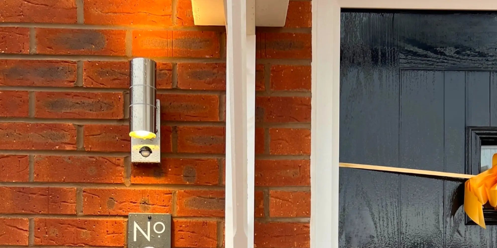 a silver wall light thats turned on, mounted on a red brick wall. the photo was taken during the day. Underneath the light is a slate sign that says 8 on it