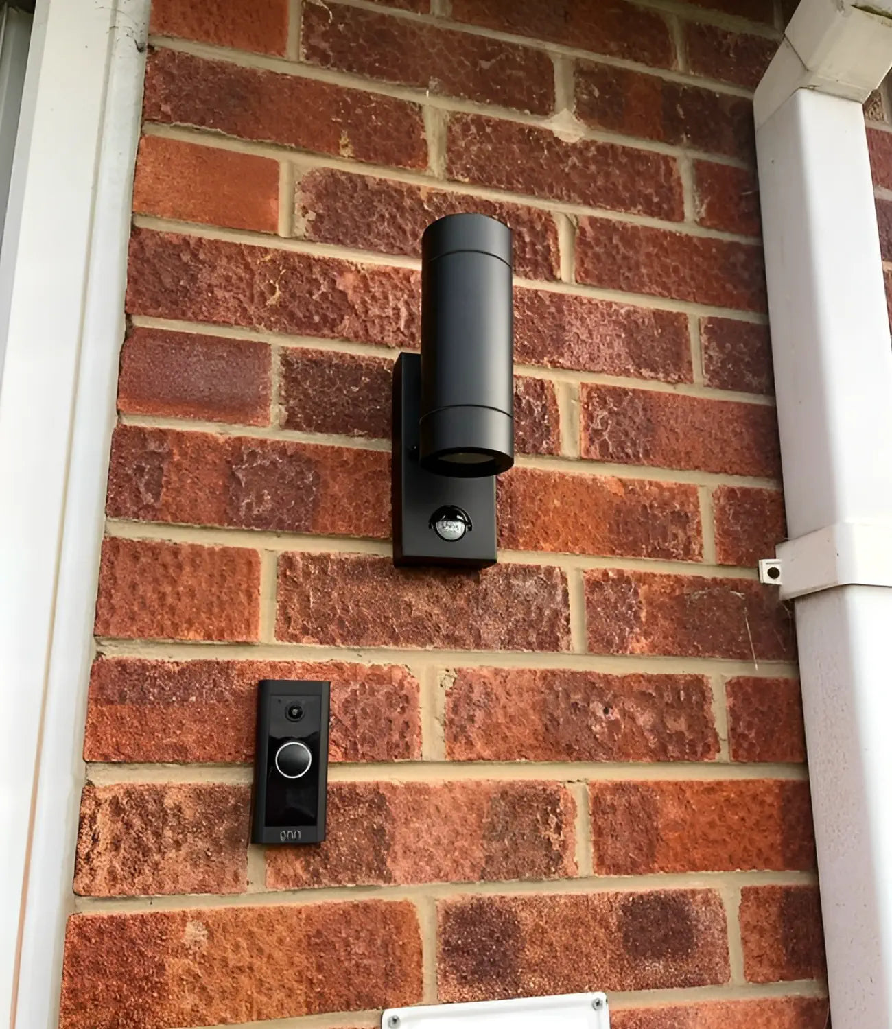 a black up and down wall light installed on a bred brick wall. There is a black ring doorbell positioned underneath and white guttering to the right.